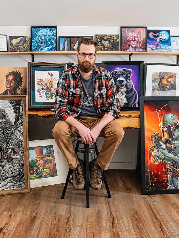 Artist with beard and glasses standing in front of multiple works of art