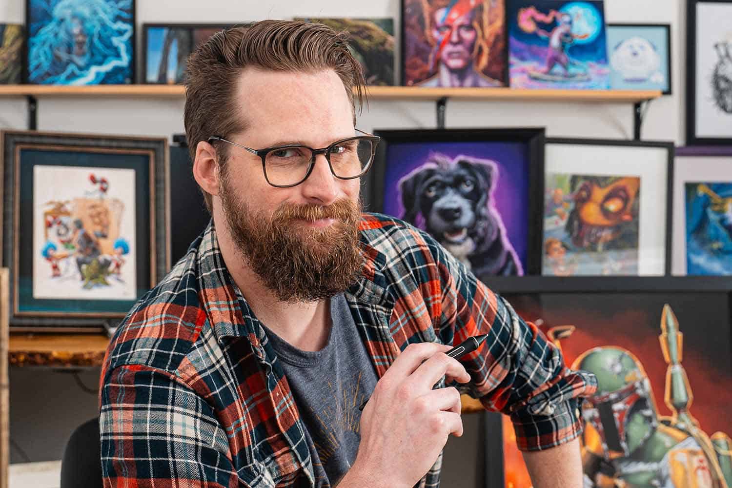 Artist wearing flannel shirt and glasses sitting in front of a wall of original paintings